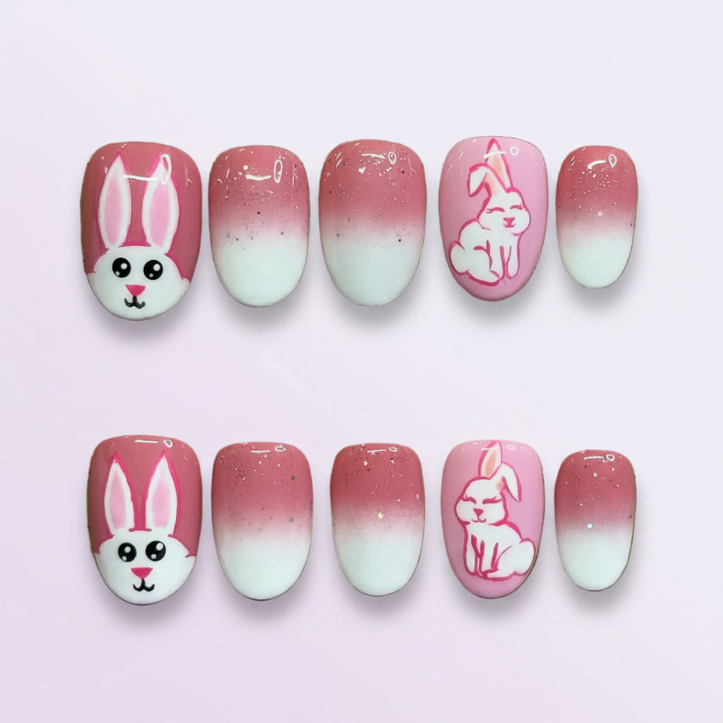 A set of 10 press-on nails displayed on a brown cardboard backing, featuring pink and white ombre designs with bunny and floral illustrations.