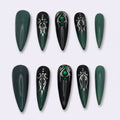 A set of ten false press-on nails arranged in two rows on a light gray background. The nails are almond-shaped, featuring a color scheme of dark forest green, black, and metallic silver gothic patterns, with two nails decorated with small green gem accents.