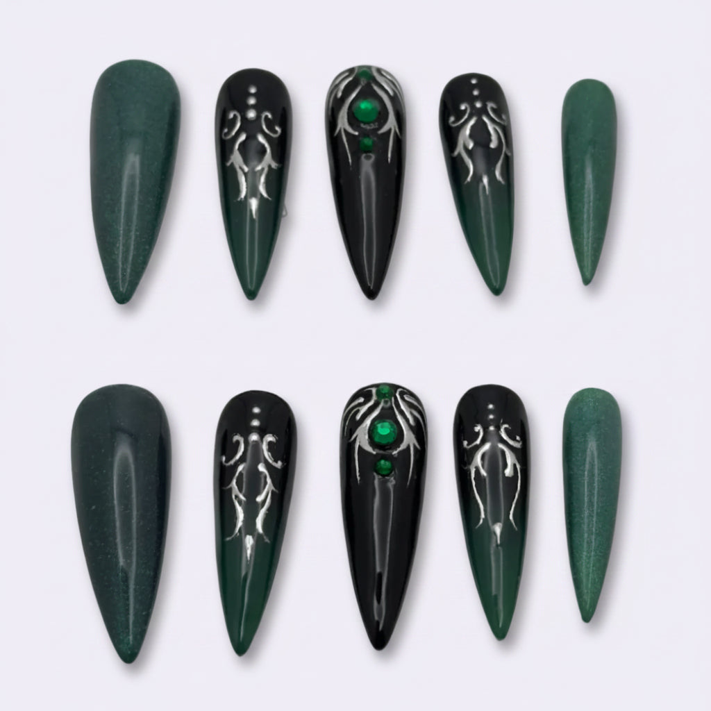 A set of ten false press-on nails arranged in two rows on a light gray background. The nails are almond-shaped, featuring a color scheme of dark forest green, black, and metallic silver gothic patterns, with two nails decorated with small green gem accents.