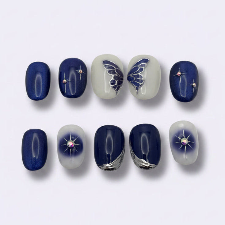A set of 10 press-on nails arranged in two rows on a light gray background. The nails feature various designs including solid deep blue, white and blue gradient nails with butterfly and star details, and dark blue nails with silver glitter and purple star accents.