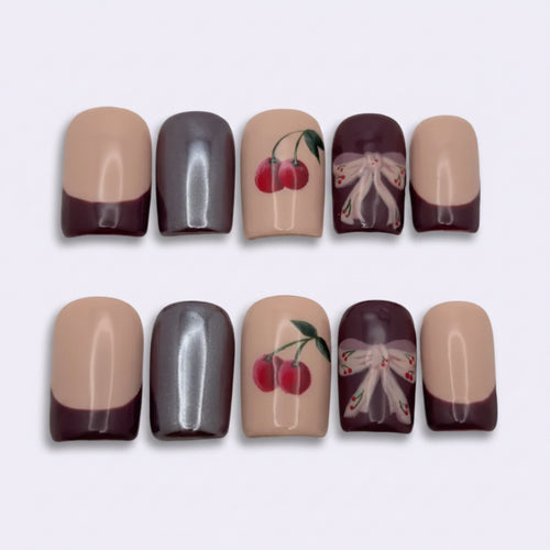 Ten false press-on nails arranged in two rows of five on a plain light background. The nails feature a two-tone design with a warm nude beige top half and a deep plum purple bottom half, with two additional nails featuring cherry and bow illustrations on the top half.