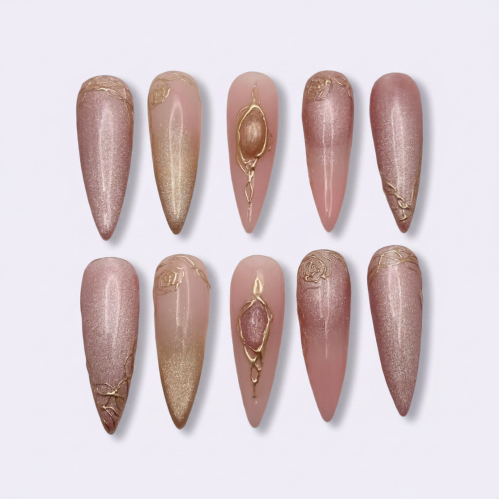 Ten stiletto-shaped press-on nails arranged in two rows on a light gray background. The nails feature a glossy metallic pink and champagne gold gradient with delicate gold glitter accents and a central rhinestone design on two of the nails.