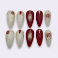 A set of ten false press-on nails arranged in two rows on a light gray background. The nails feature a glossy deep red and creamy white color scheme, decorated with gold glitter lines, rose motifs, and red jewel accents.