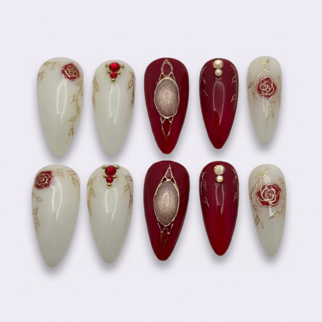 A set of ten false press-on nails arranged in two rows on a light gray background. The nails feature a glossy deep red and creamy white color scheme, decorated with gold glitter lines, rose motifs, and red jewel accents.