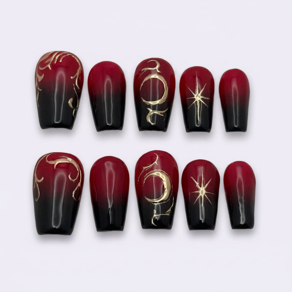 A set of ten press-on nails arranged in two rows of five on a plain light gray background. The nails feature a glossy black and deep red ombre gradient with metallic gold celestial and tribal-style designs.