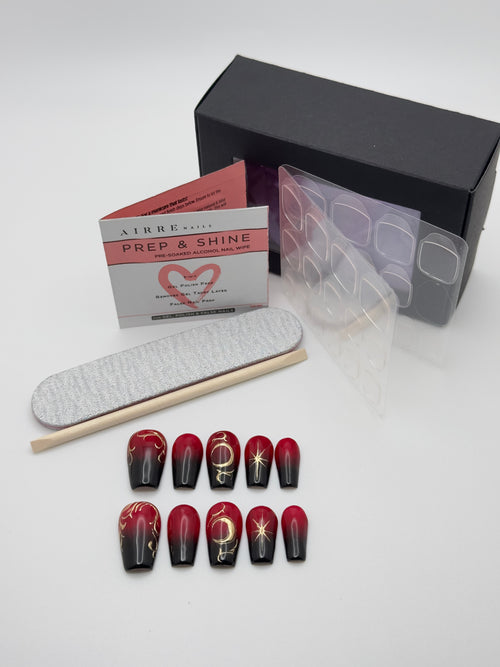 A set of red and black ombre coffin-shaped press-on nails with gold celestial designs, displayed alongside a nail file, wooden stick, clear nail stickers, and a black box on a white background.