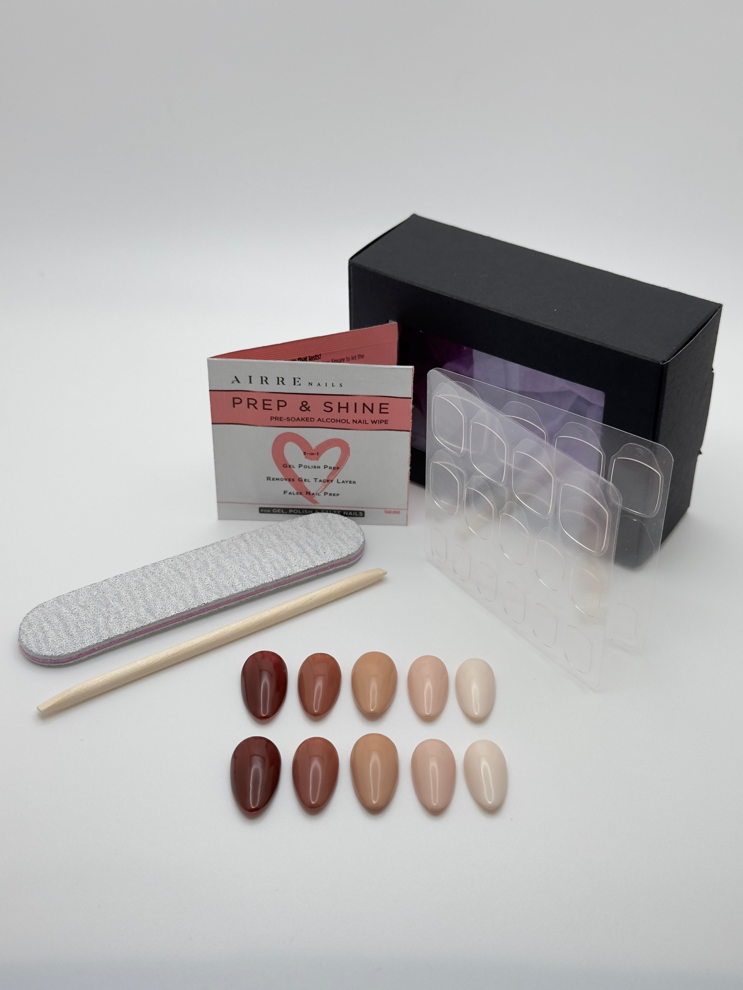 A nail kit on a light gray background, including a black box, a white and pink 'PREP & SHINE' booklet, a gray nail file, a wooden stick, a clear plastic tray, and ten false nail tips in various nude and brown shades.