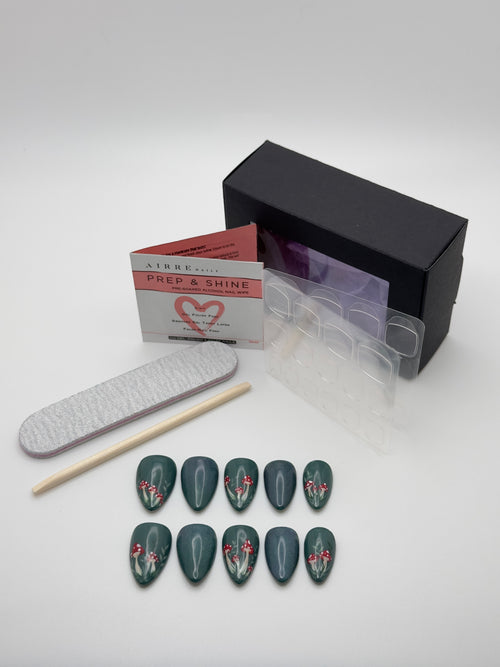 A nail art kit on a light gray background, featuring a set of dark green coffin-shaped press-on nails with white and red floral designs, a black box, a clear plastic tray, a nail file, a wooden stick, and a product card by Airbrushed.