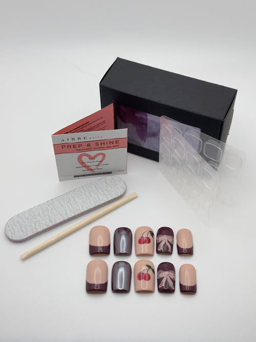 A nail art kit on a white background, including a black box, a card labeled 'PREE & SHINE', a gray nail file, a wooden stick, and ten false nails in nude, gray, and cherry blossom designs.