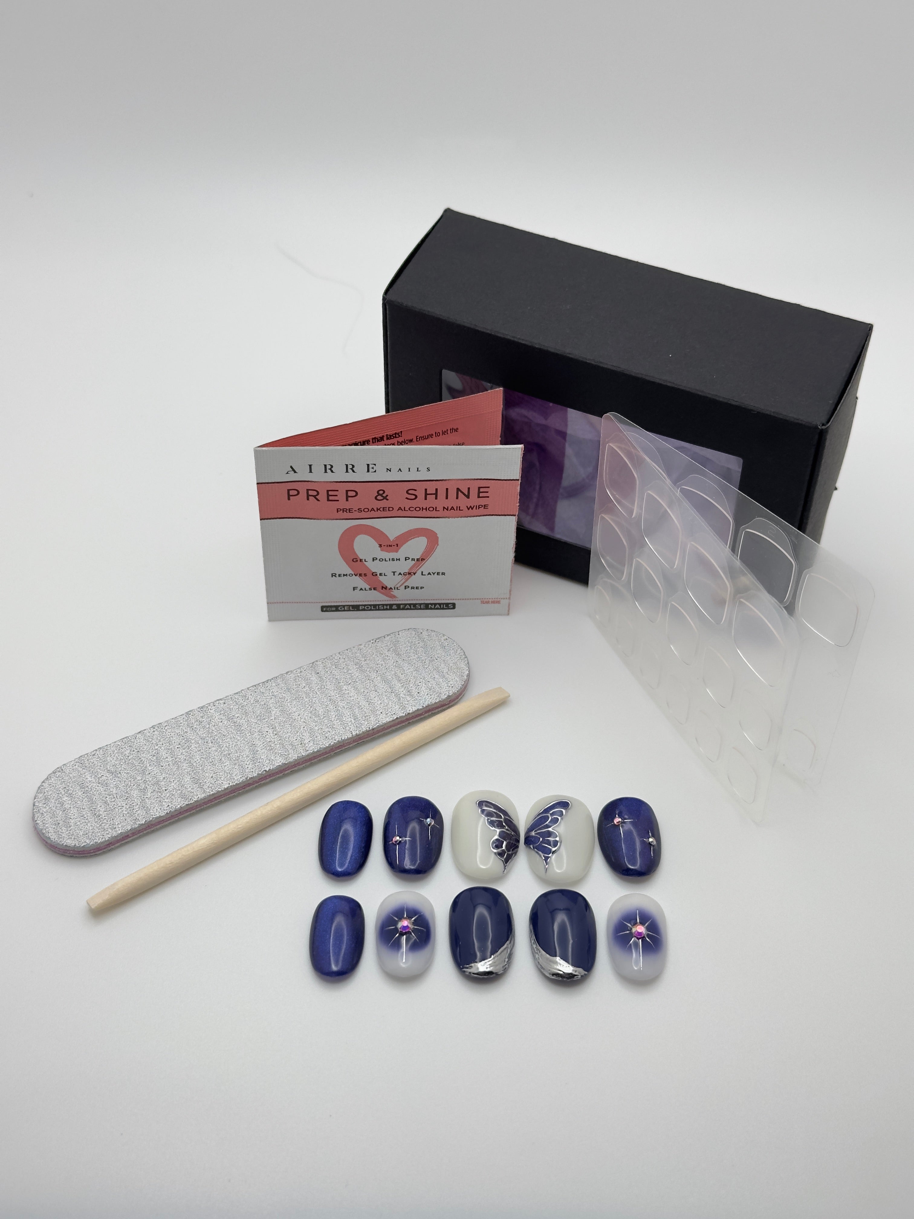 A nail art kit on a white background, including a black box, a white Prep & Shine card, a nail file, a wooden stick, clear plastic sheets, and several false nails in blue, white, and silver with decorative patterns.