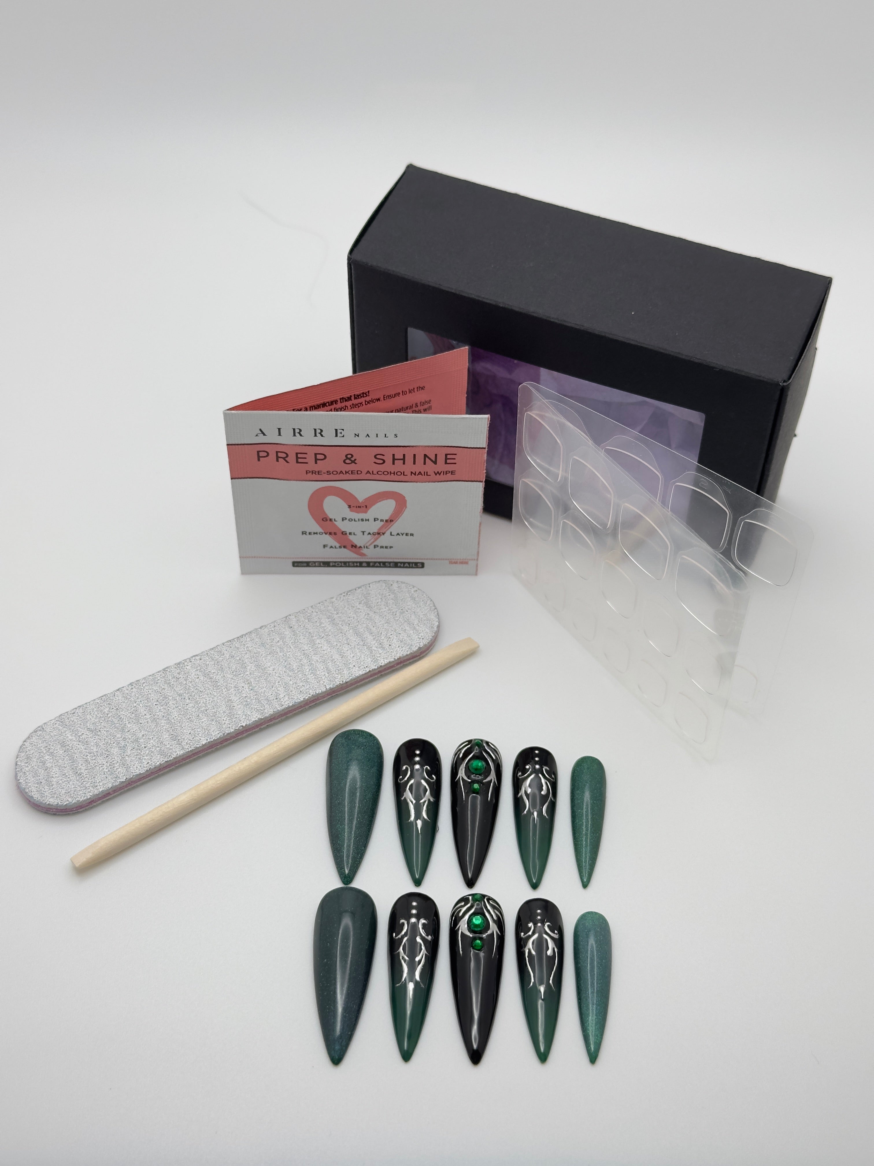 Nail art kit with dark green and black stiletto press-on nails featuring silver and green graphic designs, a white nail file, wooden cuticle stick, clear plastic sheet, and a black product box on a white background.