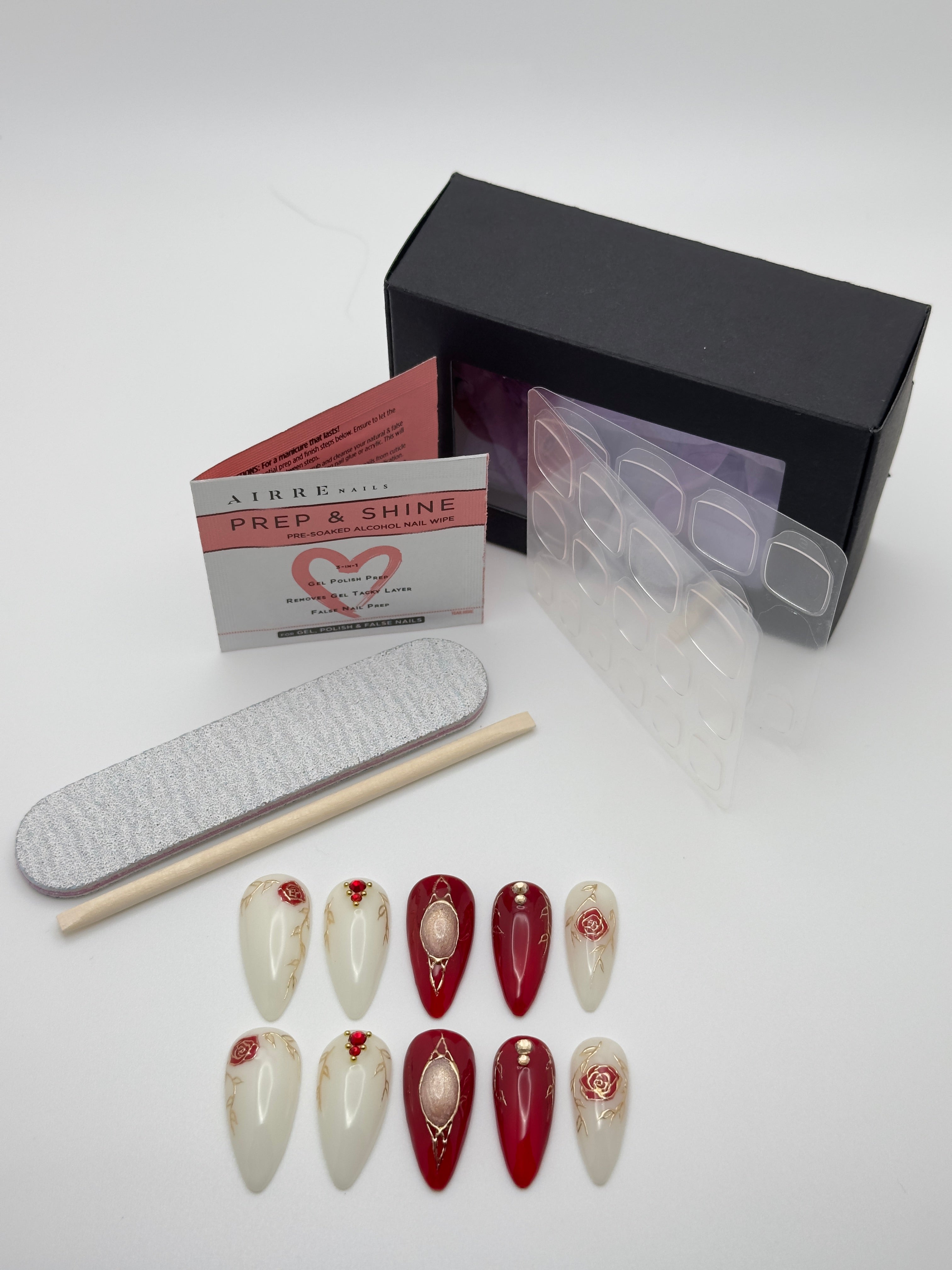 A nail art kit on a white background, including a black box, a clear tray of press-on nail templates, a gray nail file, a wooden cuticle stick, a small card labeled AFBE PREP & SHINE, and ten false nails in glossy white and deep red with floral and pearl details.