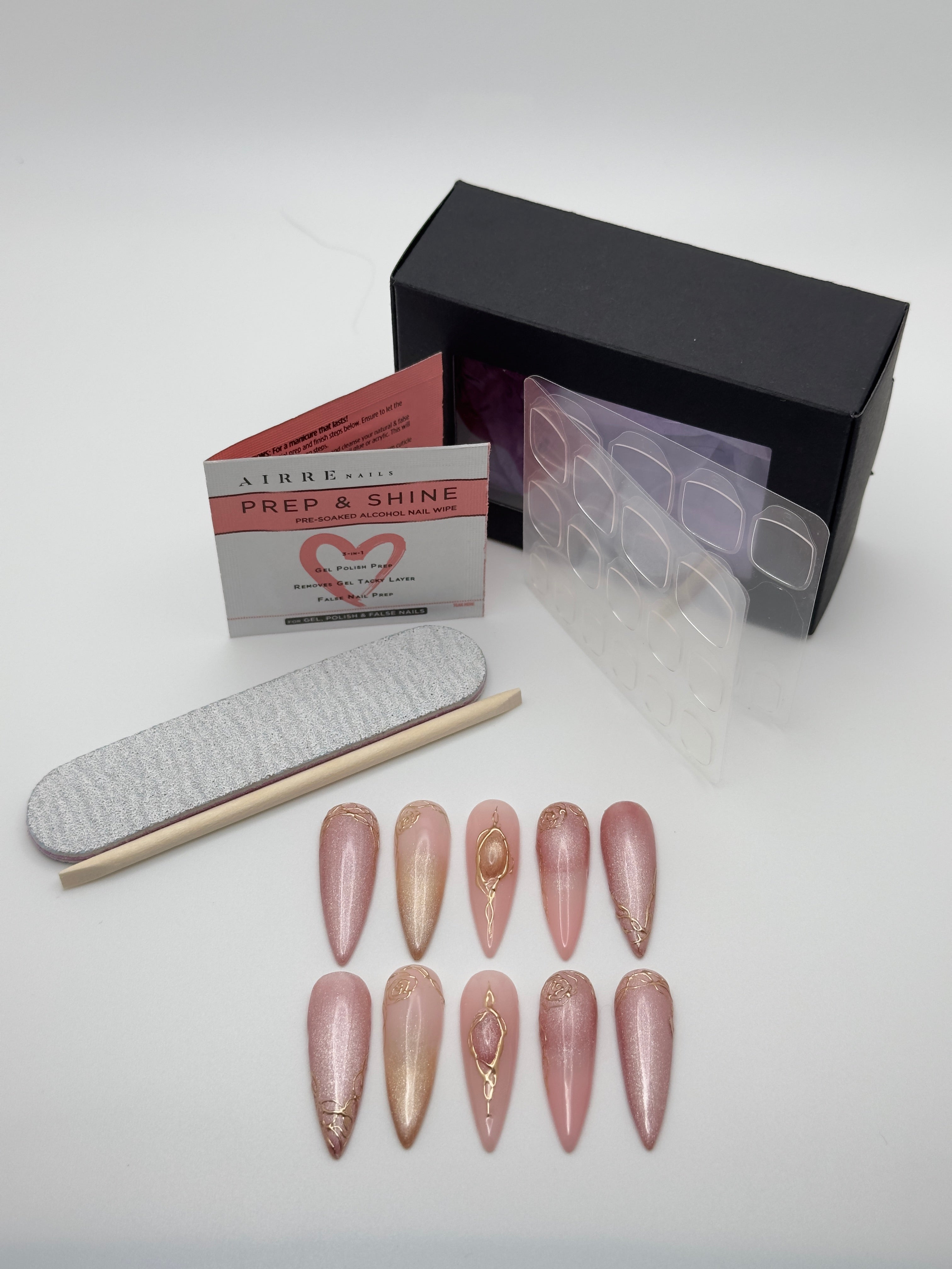 A nail art kit on a white background, featuring ten stiletto-shaped press-on nails in pink and nude shades with gold glitter and metallic designs, a gray nail file, a wooden nail stick, a clear plastic tray, a white and red instruction card, and a black box.