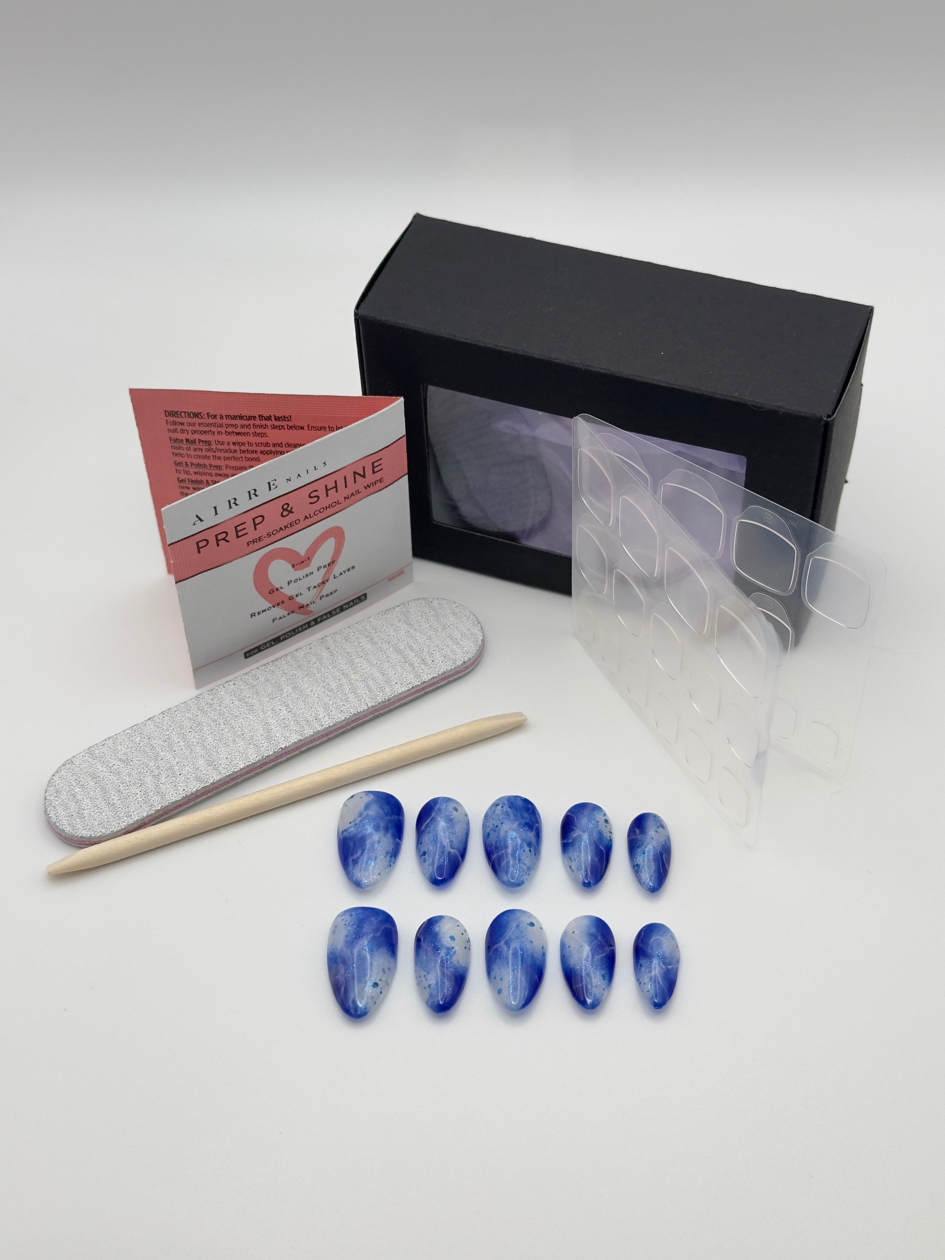 A nail kit on a light gray background, featuring blue marbled press-on nails, a black box, a clear sticker sheet, a white nail file, a wooden stick, and a red and white instruction card.