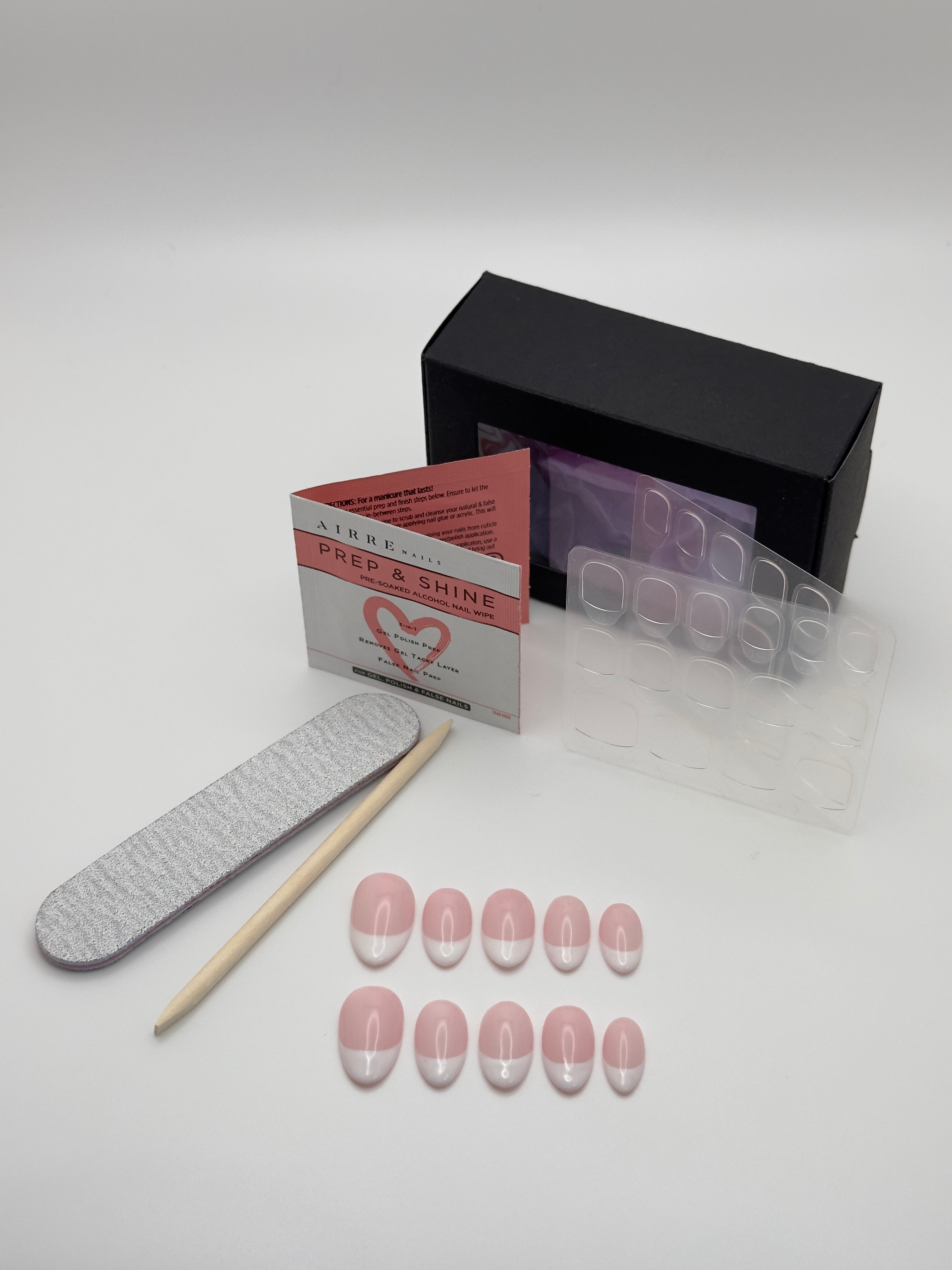 A nail art kit on a light gray background, including a black box, a clear plastic tray, pink and white nail tips, a gray nail file, a wooden stick, and a Prep & Shine card.